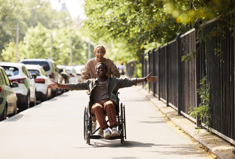 Happy excited young African-American disabled man in wheelchair pushed by running woman outstretching arms and feeling freedom
